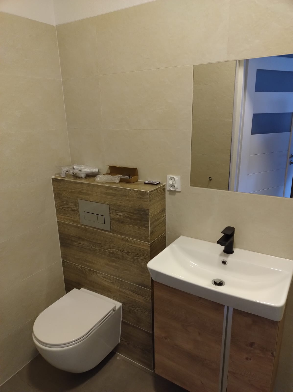 Nowoczesna łazienka z beżowymi płytkami i drewnopodobną zabudową WC oraz umywalką z czarną armaturą. Widoczne akcesoria instalacyjne.
