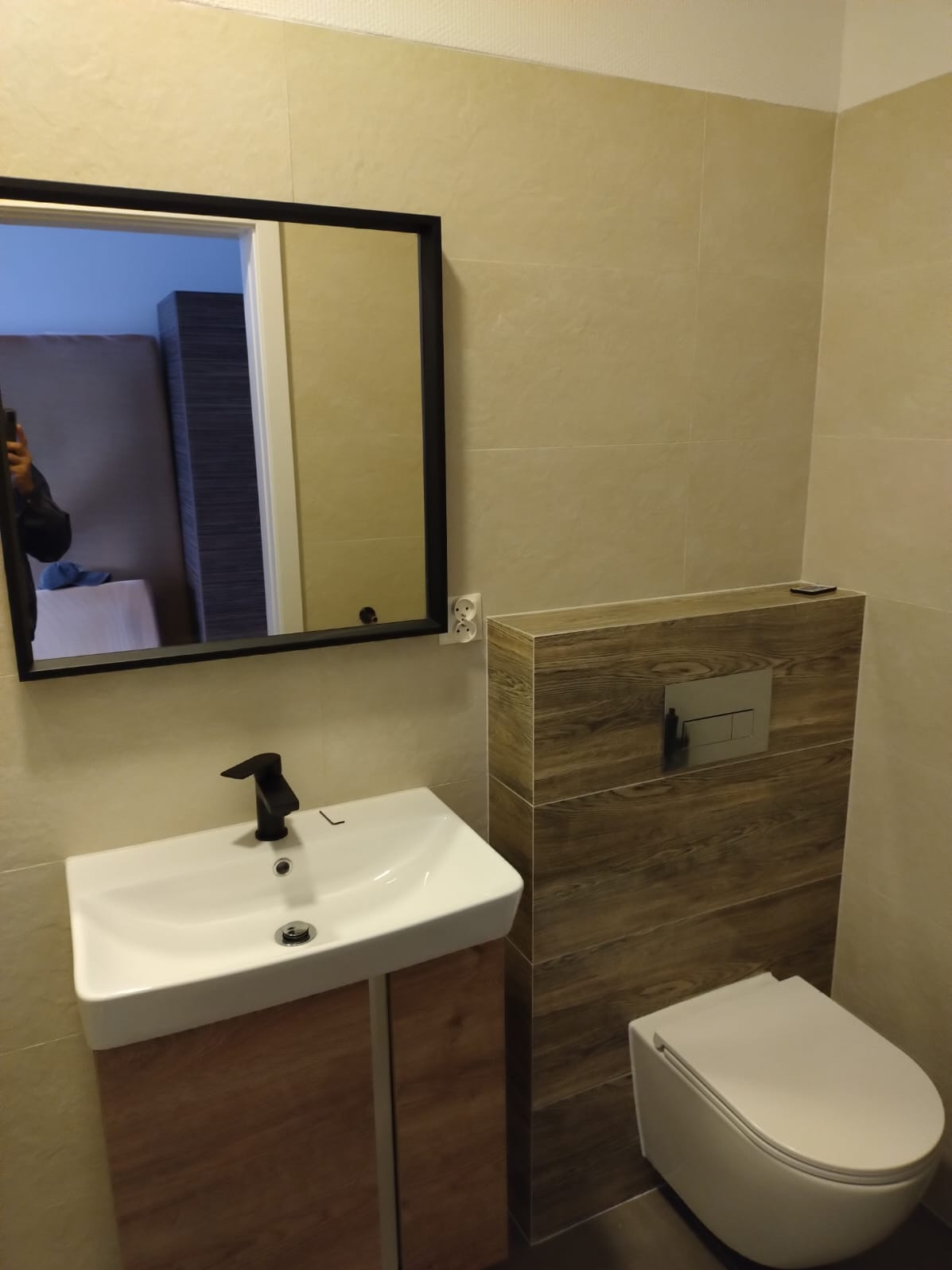 Nowoczesna łazienka z beżowymi płytkami, umywalką na szafce z drewnopodobnym frontem, czarną armaturą i wiszącą toaletą z obudową z płytek drewnopodobnych.