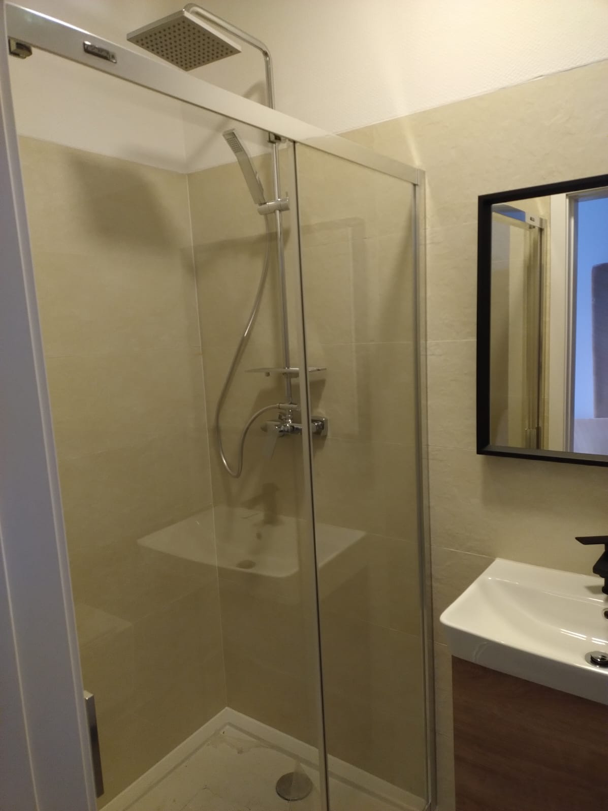 Nowoczesna kabina prysznicowa ze szklanymi drzwiami przesuwnymi i chromowaną armaturą. Beżowe płytki ścienne i umywalka z szafką w odcieniach brązu.