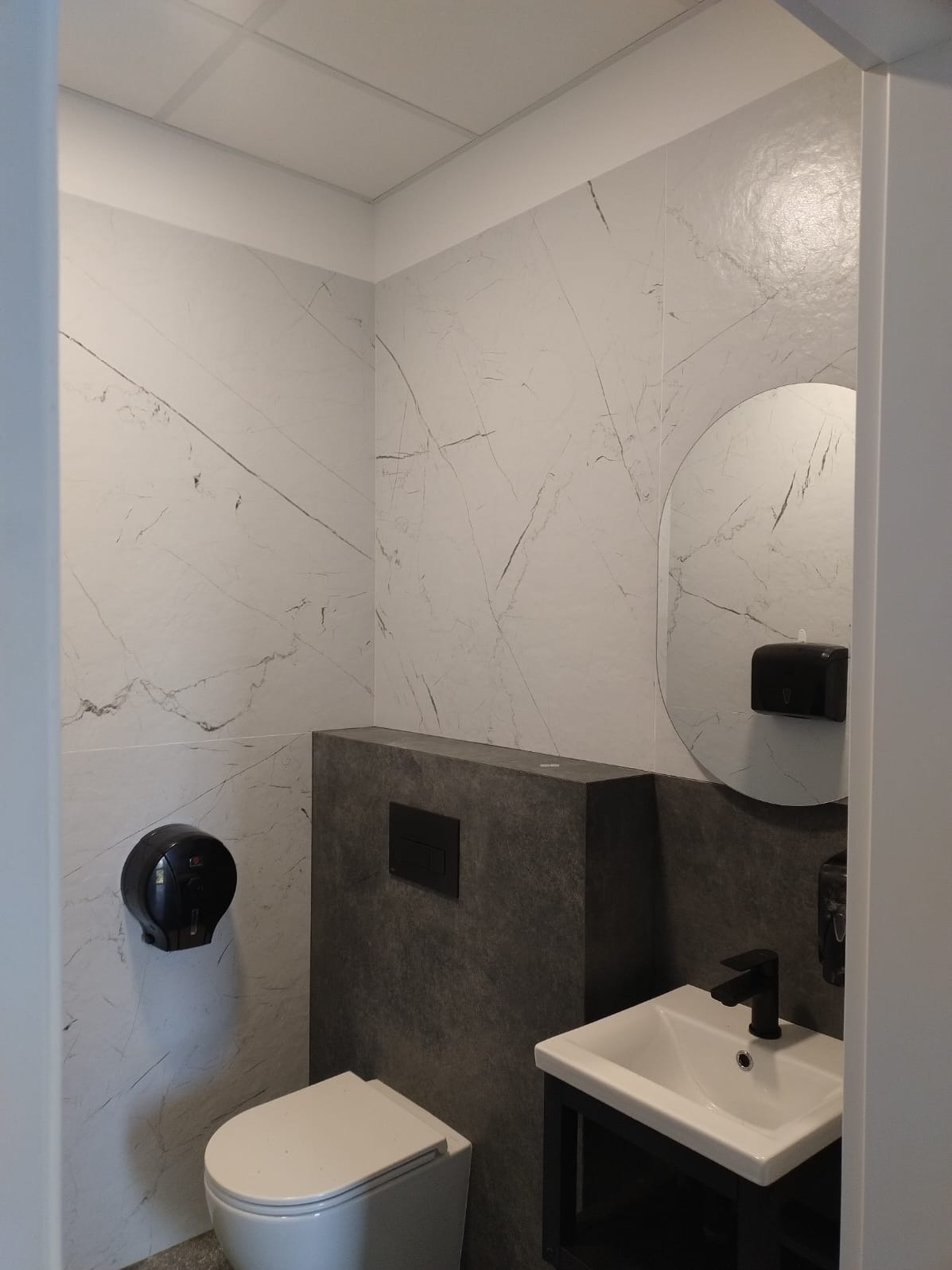 Nowoczesna łazienka z marmurowymi płytkami, okrągłym lustrem i czarną armaturą. Umywalka nablatowa na czarnym stelażu, wisząca toaleta. Minimalistyczny design.