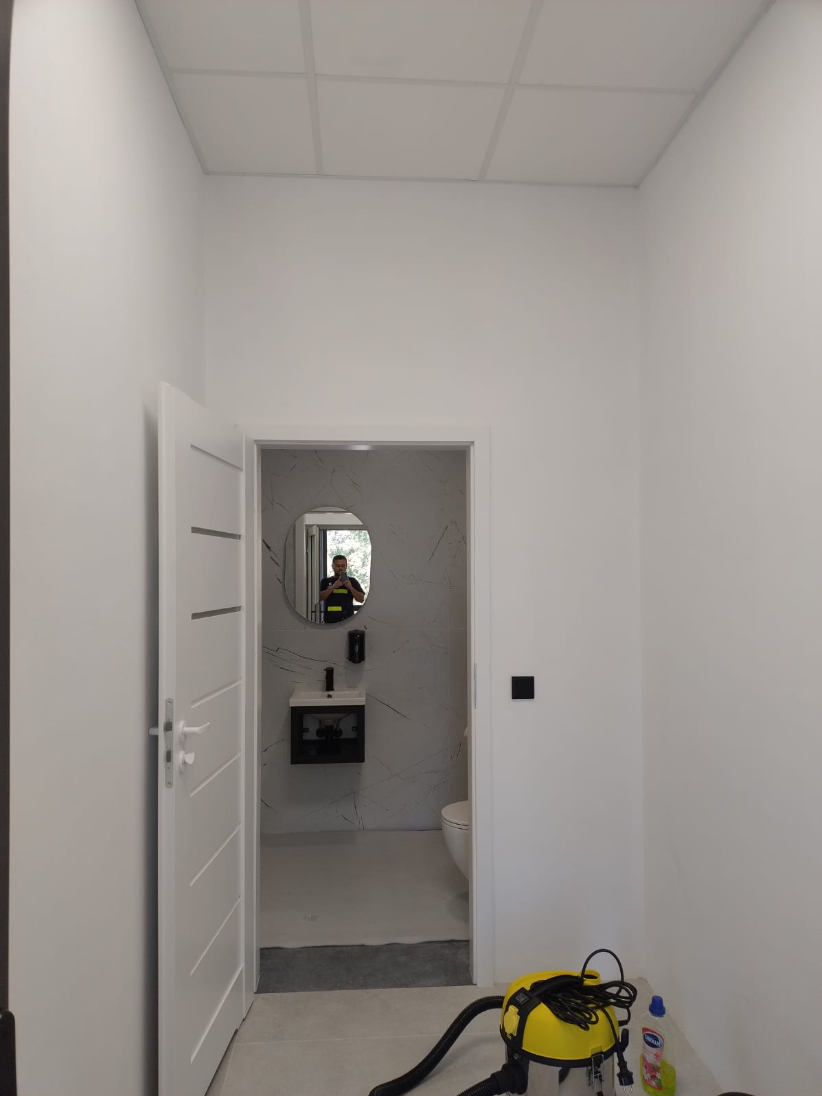 Białe wnętrze z otwartymi drzwiami do łazienki po remoncie. Widoczne umywalka, lustro, płytki imitujące marmur i odkurzacz. Na ścianie włącznik światła.