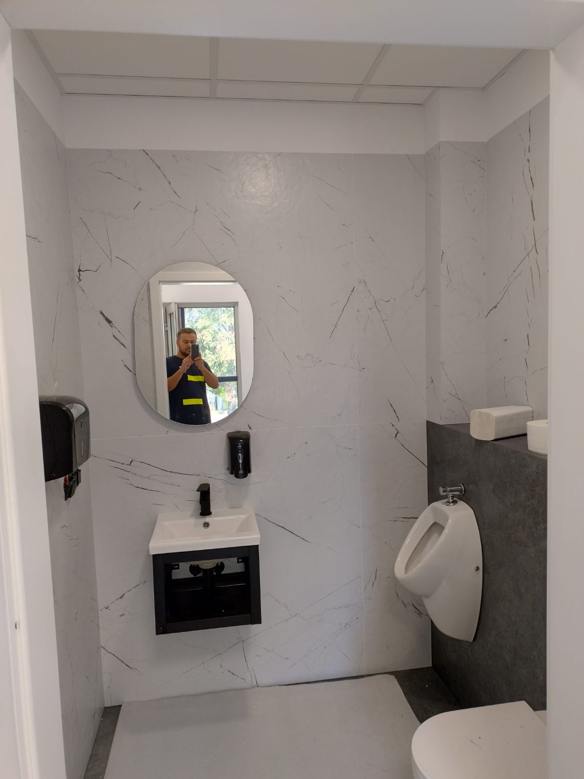 Wykończona łazienka z umywalką, pisuar i lustrem owalnym, w którym odbija się osoba robiąca zdjęcie. Ściany wyłożone płytkami imitującymi marmur.