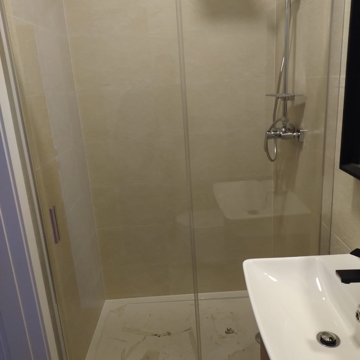 Beżowa łazienka po remoncie: kabina prysznicowa ze szklanymi drzwiami, chromowana armatura i biała umywalka. Minimalistyczny design z jasnymi płytkami.