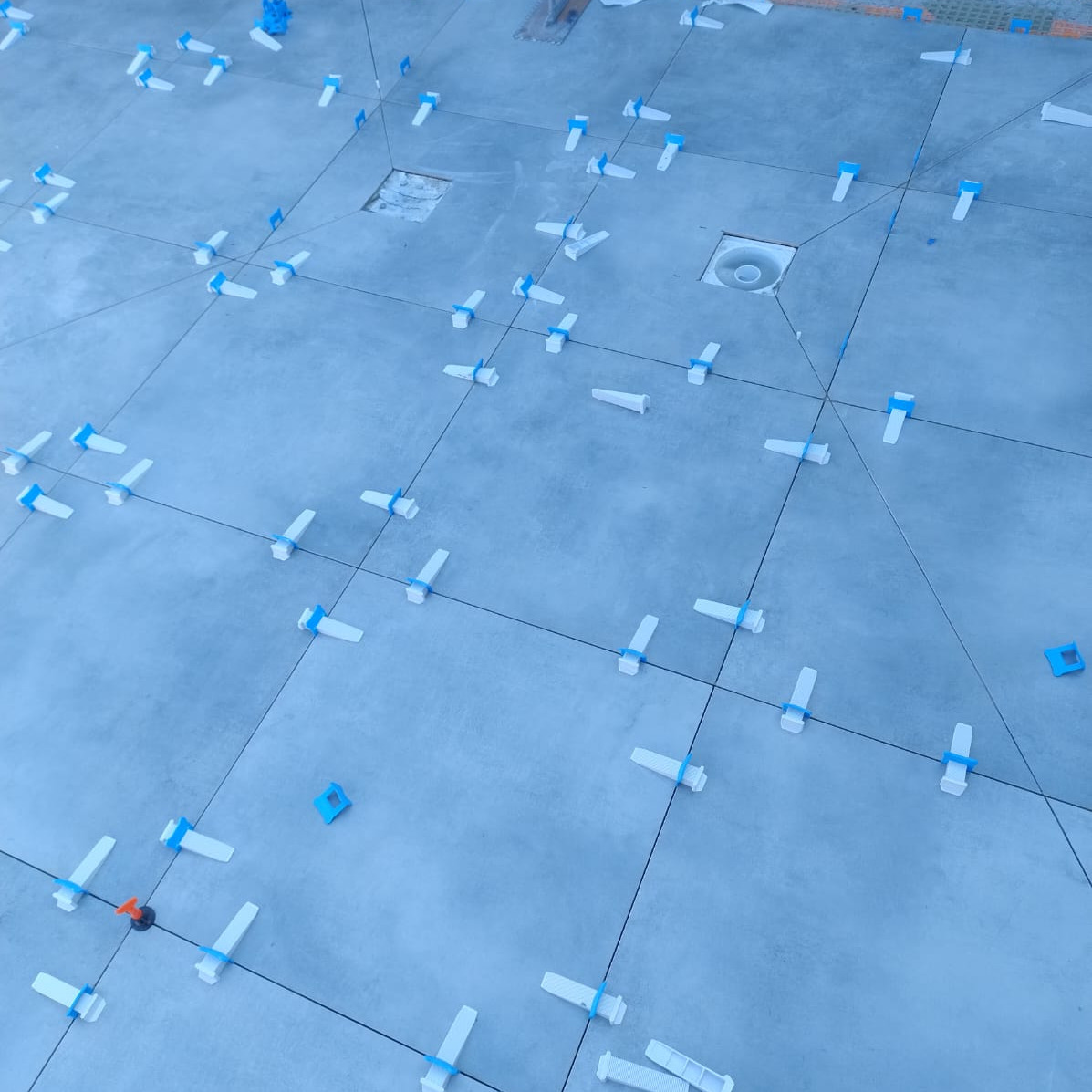 Ułożone szare płytki podłogowe z systemem poziomowania, widoczne fugi i odpływ liniowy. Etap prac glazurniczych, bliska perspektywa.