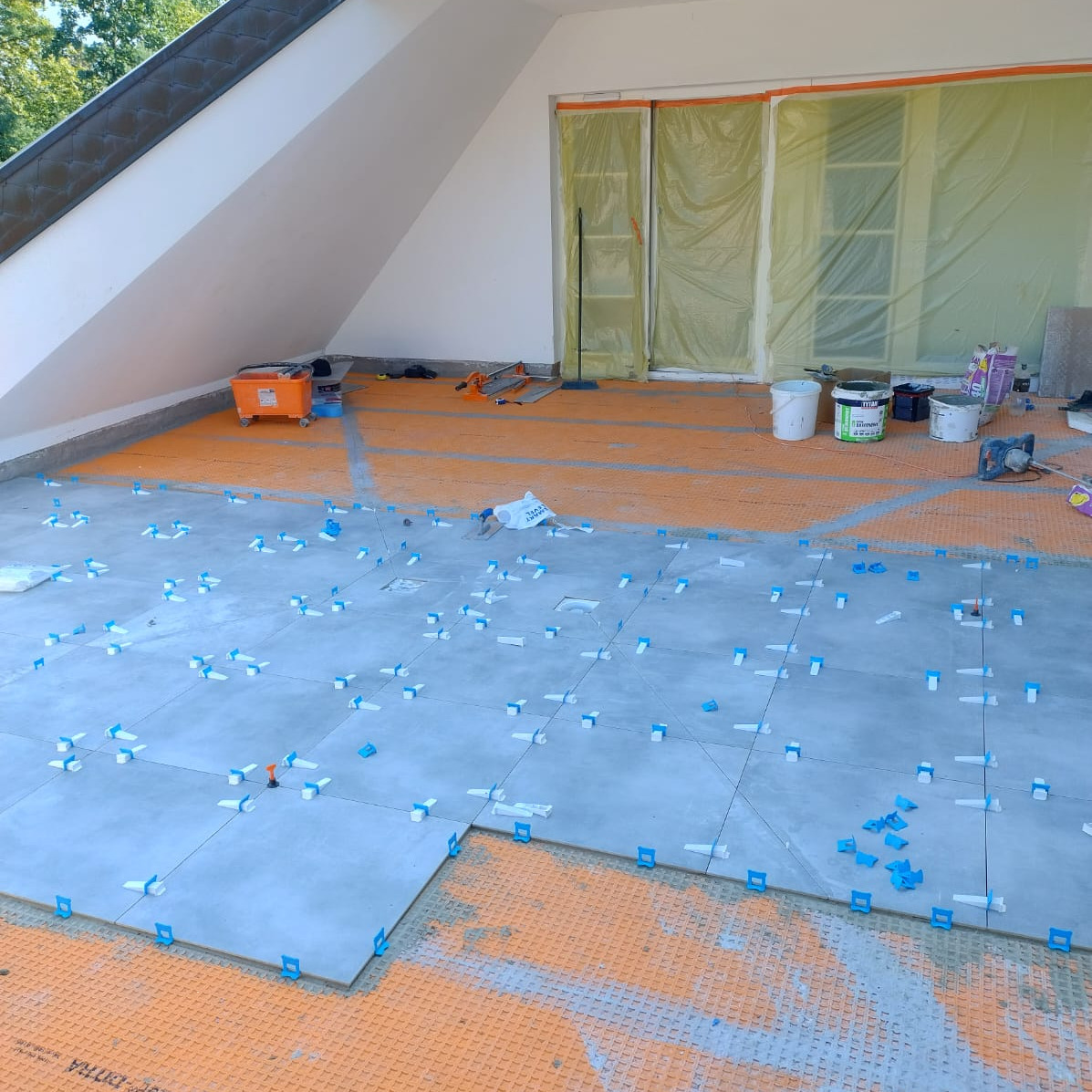 Układanie szarych płytek z użyciem niebieskich klipsów poziomujących na pomarańczowej macie uszczelniającej w jasnym, skośnym pomieszczeniu.