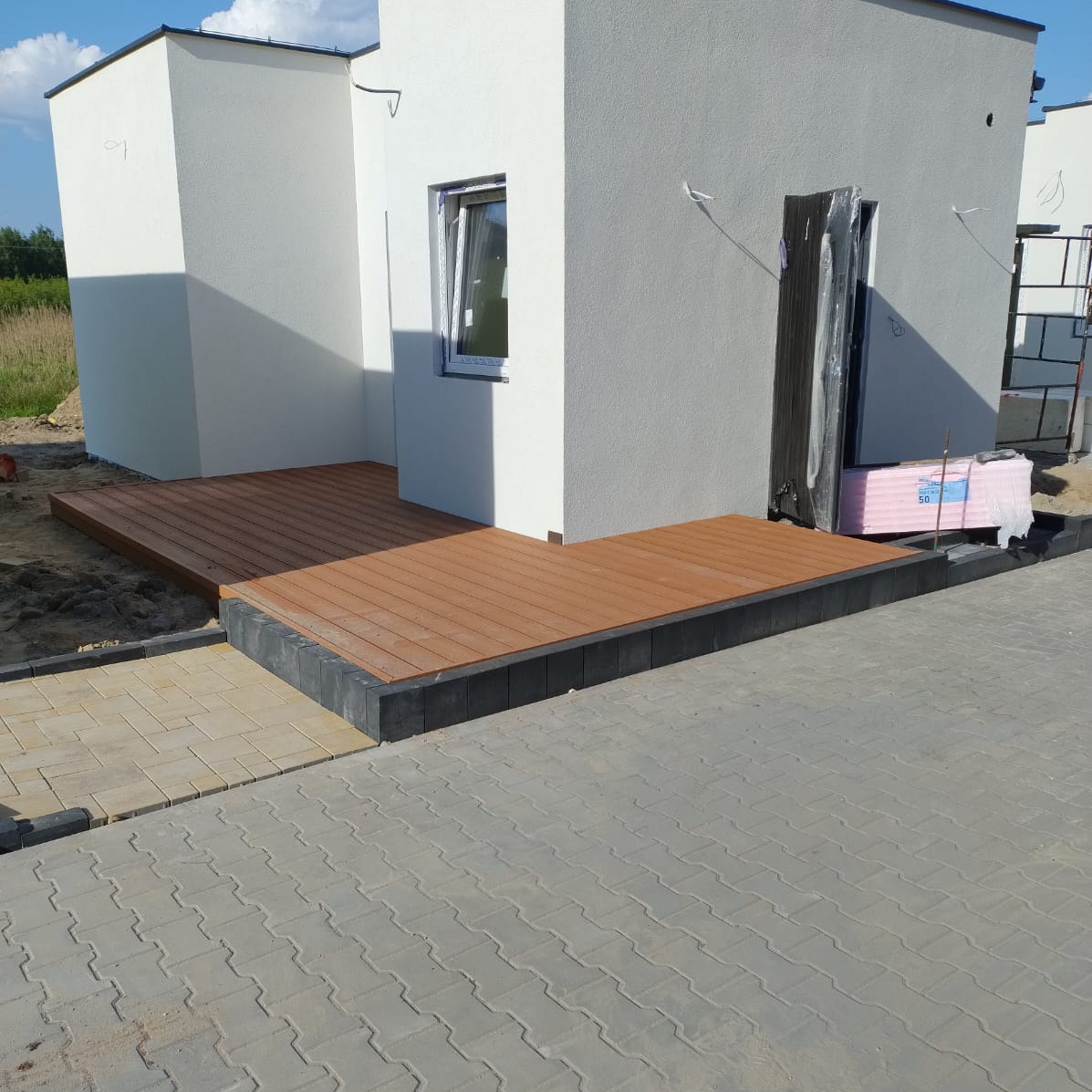 Nowoczesny taras z desek kompozytowych przy białym budynku, z kostką brukową i elementami wykończeniowymi. Widok z zewnątrz, w słoneczny dzień.