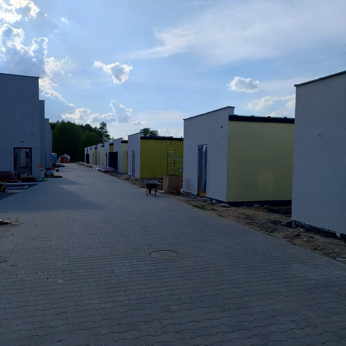 Szereg budynków w trakcie wykończenia na osiedlu, z widocznymi rusztowaniami i materiałami budowlanymi. Część budynków pomalowana na żółto, inne w szarym tynku.