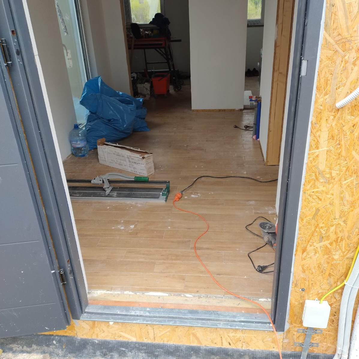 Wnętrze w trakcie prac glazurniczych: ułożone płytki podłogowe imitujące drewno, narzędzia glazurnicze, worki budowlane, widok przez otwarte drzwi.