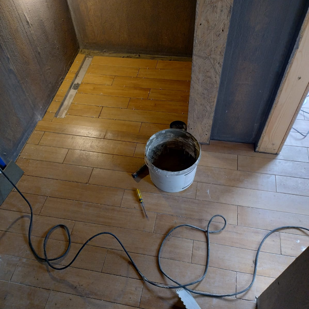Układanie drewnopodobnych płytek w łazience. Widoczny odpływ liniowy, wiadro z klejem i kabel. Etap prac wykończeniowych w trakcie remontu łazienki.
