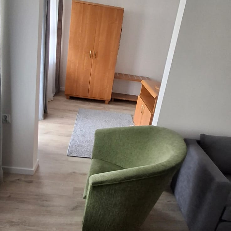 Umeblowane wnętrze pokoju z drewnianą szafą, komodą i zielonym fotelem. Jasna podłoga, szary dywan i zasłony w tle. Ujęcie z niskiego kąta.