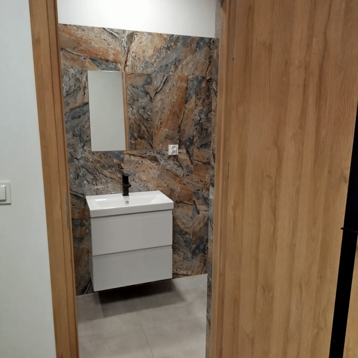 Wykończona łazienka z umywalką na szafce, lustrem i płytkami imitującymi kamień. Widok zza drewnianych drzwi, z szarą podłogą. Gniazdko elektryczne widoczne na ścianie.