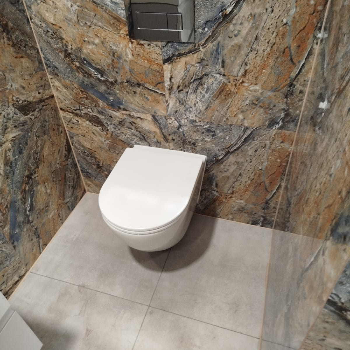 Nowoczesna toaleta podwieszana z szarą płytką podłogową i dekoracyjną okładziną ścienną imitującą marmur w odcieniach brązu i szarości.