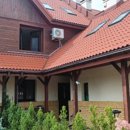 Adam Szarałło Klima - Drewniana elewacja domu z białą jednostką zewnętrzną klimatyzacji obok otwartego okna, czerwony dach z oknami dachowymi i zadaszony taras z ceglaną podmurówką.