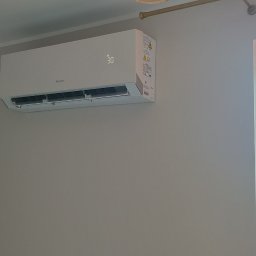 Adam Szarałło Klima - Biała jednostka klimatyzacji GREE z cyfrowym wyświetlaczem '30' na jasnoszarej ścianie, obok okna z widokiem na zieleń, czerwony dach i tory kolejowe.
