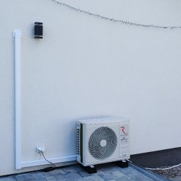 Adam Szarałło Klima - Zewnętrzna jednostka klimatyzatora Rotenso na czarnych podstawkach przy białej ścianie domu, z widoczną maskownicą instalacji i oknem tarasowym. W tle zielony ogród.