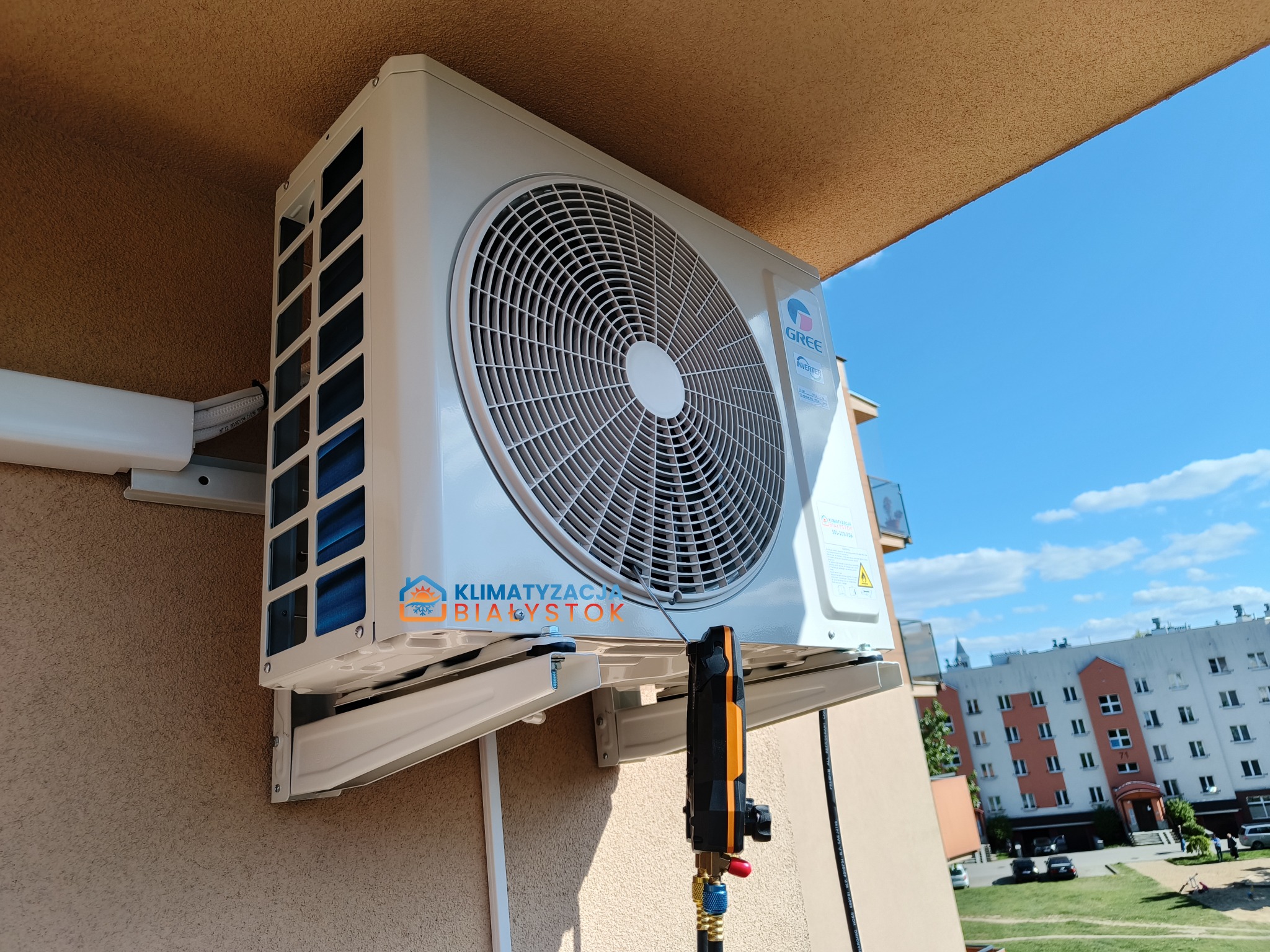 Biała jednostka zewnętrzna klimatyzacji Gree zamontowana na ścianie budynku, widoczne narzędzie pomiarowe, błękitne niebo w tle.