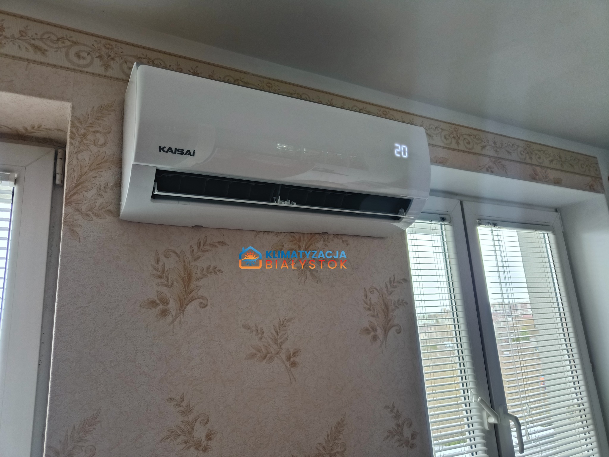 Biały klimatyzator Kaisai zamontowany na ścianie z wzorzystą tapetą, obok okna z żaluzjami. Na urządzeniu wyświetlana temperatura 20 stopni Celsjusza.