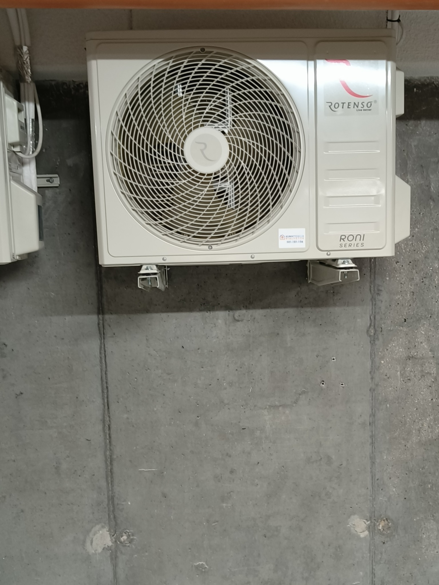 Zamontowana jednostka zewnętrzna klimatyzacji Rotenso Roni Series na szarej, betonowej ścianie. Widoczne przewody i elementy montażowe.