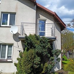 Damian Dwojakowski - Balkon z nierdzewną balustradą w domu jednorodzinnym, z satelitą na ścianie i ozdobnymi krzewami w ogrodzie. Ujęcie w słoneczny dzień.