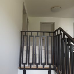 Damian Dwojakowski - Metalowa balustrada schodowa w nowoczesnym stylu, z geometrycznym wzorem. Widoczne jasne ściany i drzwi w tle. Ciemna kolorystyka balustrady kontrastuje z bielą otoczenia.