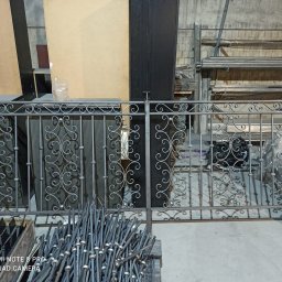 Damian Dwojakowski - Element kutej balustrady z dekoracyjnymi zawijasami, w szarym kolorze, widoczny na tle materiałów i narzędzi w warsztacie.