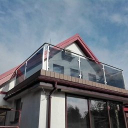 Damian Dwojakowski - Szklana balustrada balkonowa z elementami ze stali nierdzewnej na tle błękitnego nieba i dachu domu z czerwonej dachówki. Widok z dołu.