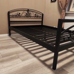 Damian Dwojakowski - Czarna, metalowa rama łóżka z dekoracyjnym zagłówkiem, widoczna na tle beżowej ściany i drewnianej podłogi. Uwagę przyciąga ornamentyka zagłówka i geometryczny wzór ramy.