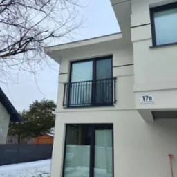 Nowoczesny budynek z balkonem zabezpieczonym czarną, metalową balustradą. Widoczny fragment elewacji, okna z żaluzjami i łopata do śniegu oparta o ścianę.