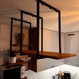 Damian Dwojakowski - Nowoczesna, metalowa konstrukcja podwieszana w kuchni, z drewnianymi półkami. Minimalistyczny design, czarne profile, ciepłe oświetlenie. Widok na kuchenny blat i sprzęty.