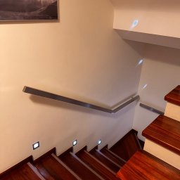 Damian Dwojakowski - Drewniane schody z metalową poręczą i oświetleniem punktowym w ścianie. Na ścianie wisi obraz z motywem górskim. Ujęcie z góry, podkreślające detale wykończenia.