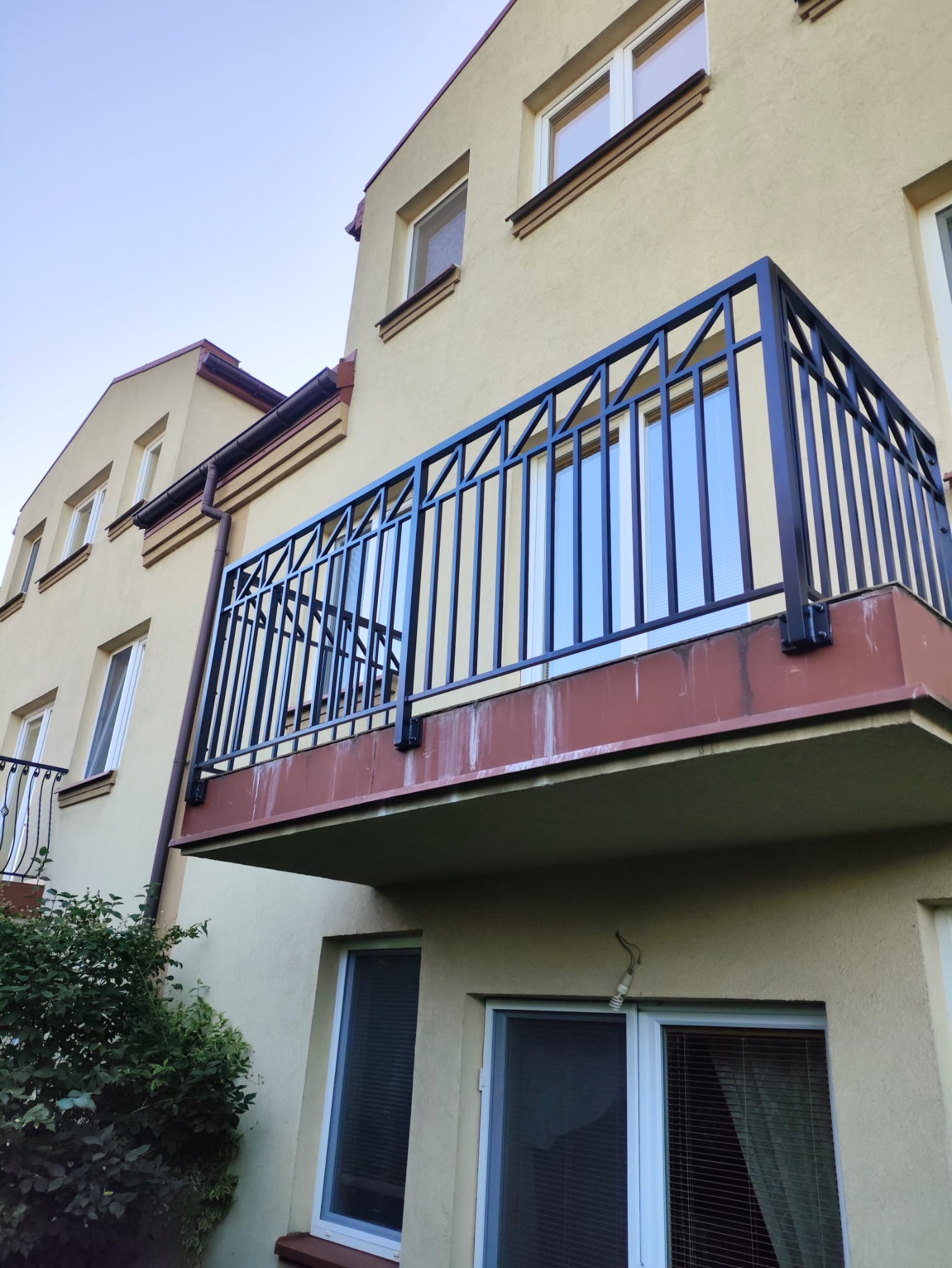 Czarna, metalowa balustrada balkonowa z pionowymi prętami, zamontowana na balkonie budynku o beżowej elewacji. Widok z dołu, zieleń w tle.