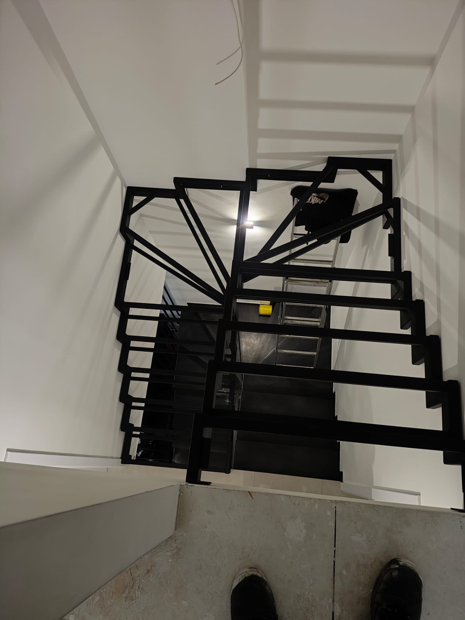 Nowoczesne, czarne schody metalowe spiralne, widziane z góry. Minimalistyczny design, białe ściany i jasna podłoga. Widoczne elementy konstrukcyjne i schody w trakcie montażu.