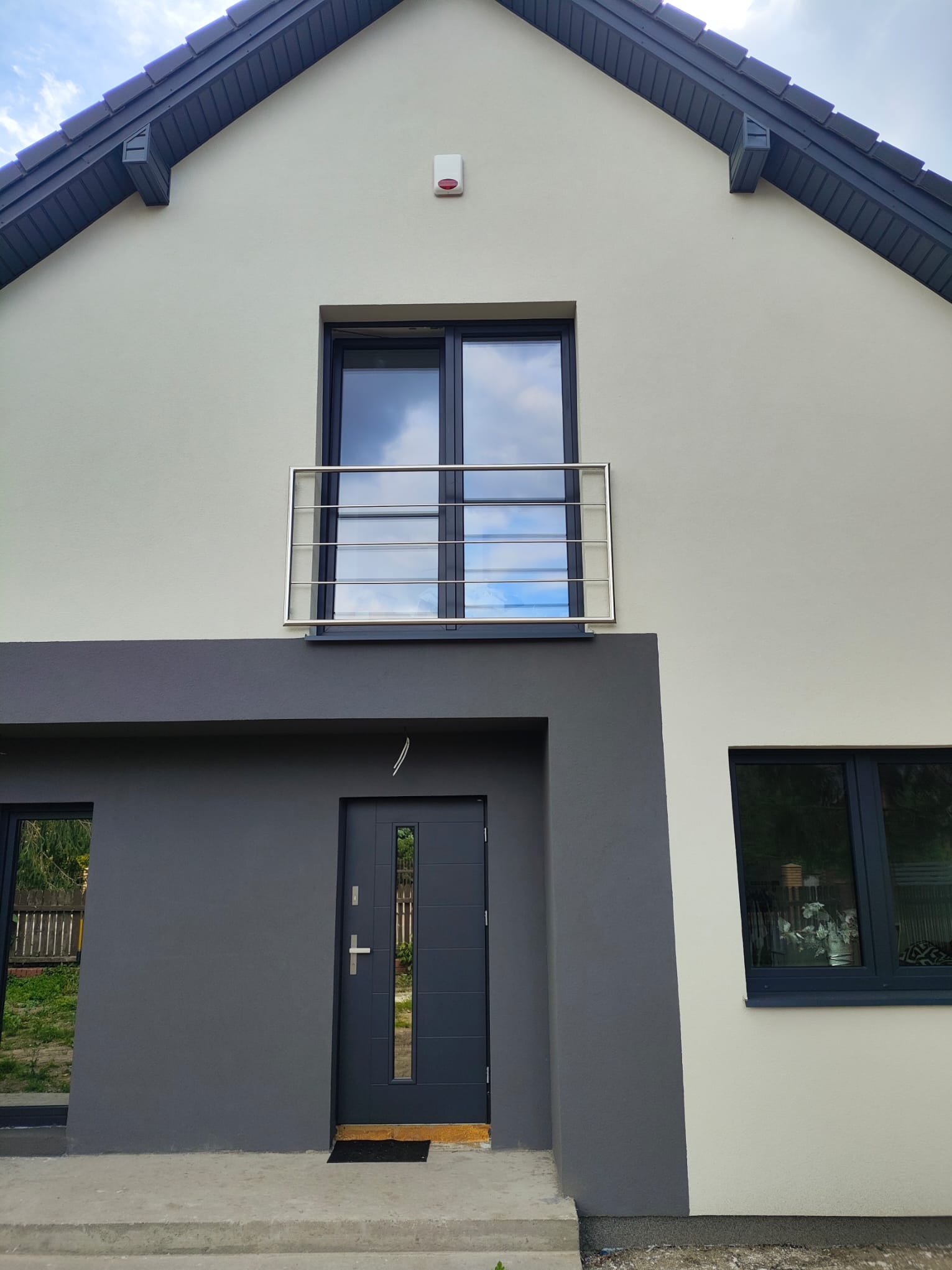 Dwukondygnacyjny dom z szarą stolarką okienną i balkonem z prostą, metalową balustradą. Elewacja w odcieniach bieli i szarości. Widok z dołu.