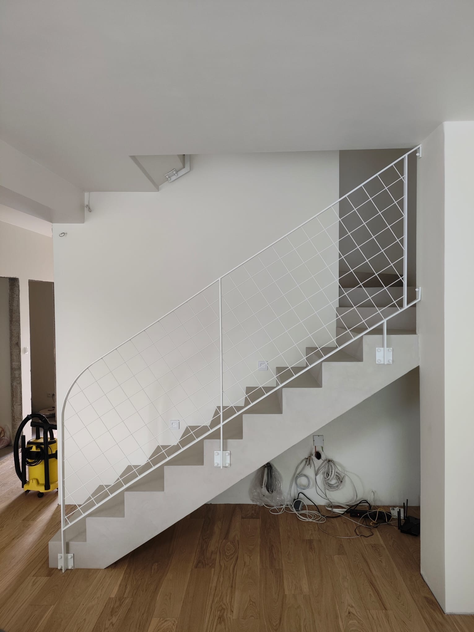 Minimalistyczne schody z białą, metalową balustradą o siatkowym wzorze. Widoczne kable pod schodami i odkurzacz w tle. Nowoczesne wykończenie wnętrza.