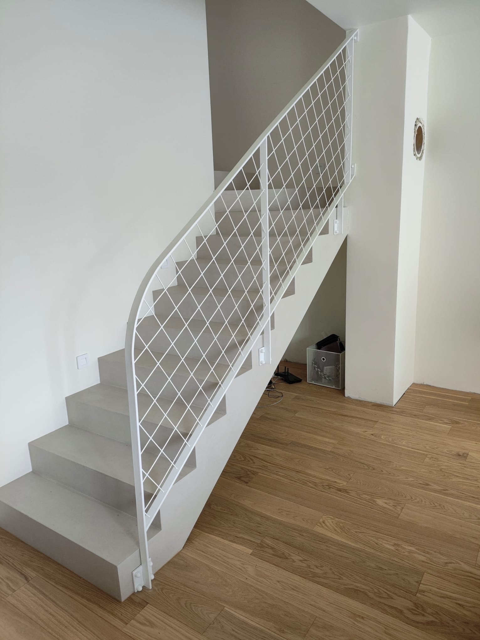 Nowoczesne schody betonowe z białą, metalową poręczą o siatkowym wzorze. Minimalistyczny design, drewniana podłoga, jasne wnętrze. Widok z dołu.