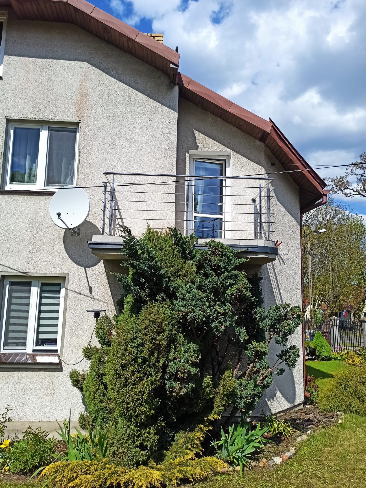 Balkon z nierdzewną balustradą w domu jednorodzinnym, z satelitą na ścianie i ozdobnymi krzewami w ogrodzie. Ujęcie w słoneczny dzień.