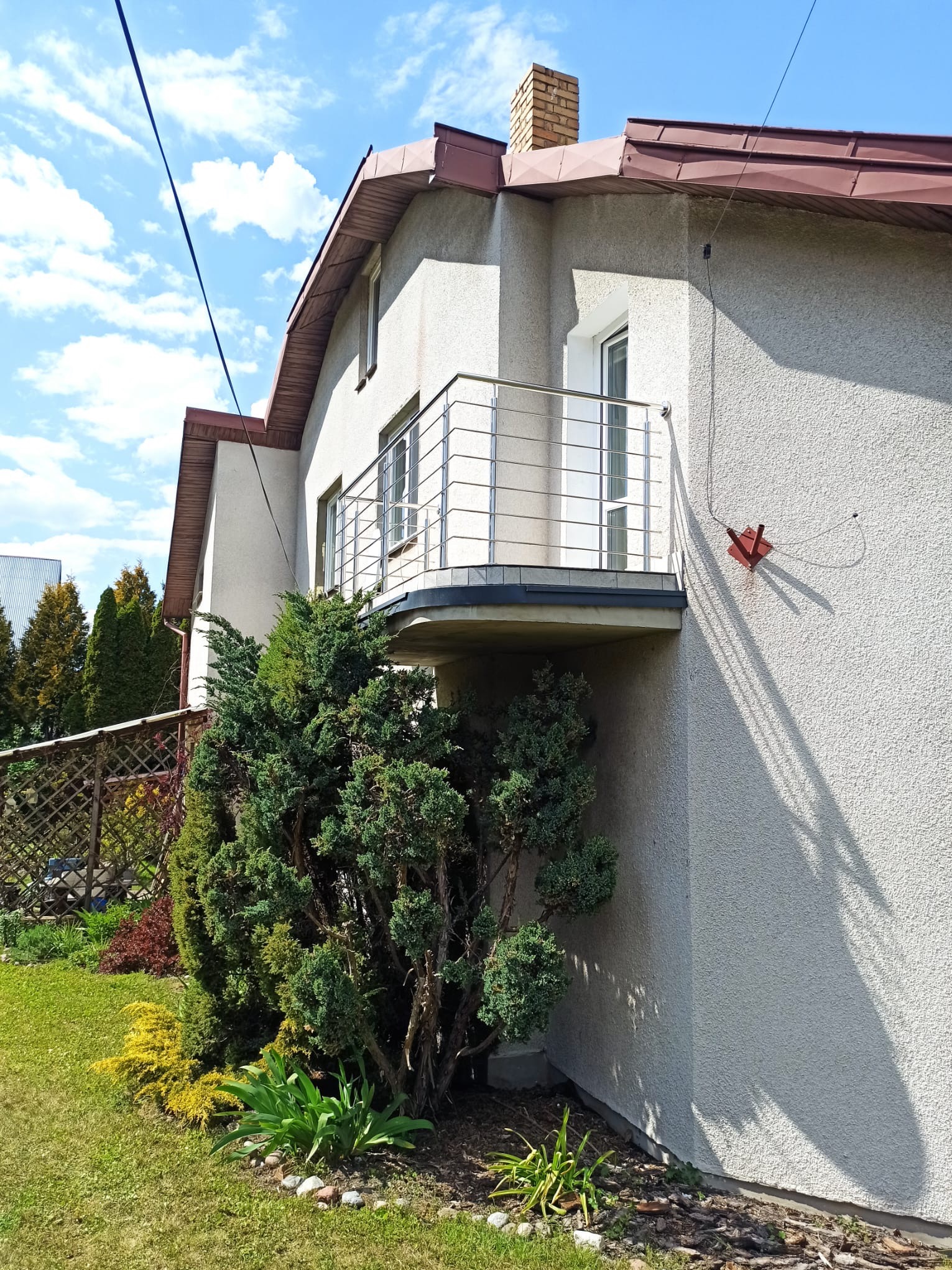 Biały dom z balkonem z balustradą ze stali nierdzewnej. Widoczne drzewa i krzewy. Błękitne niebo z chmurami. Czerwona antena na ścianie budynku.
