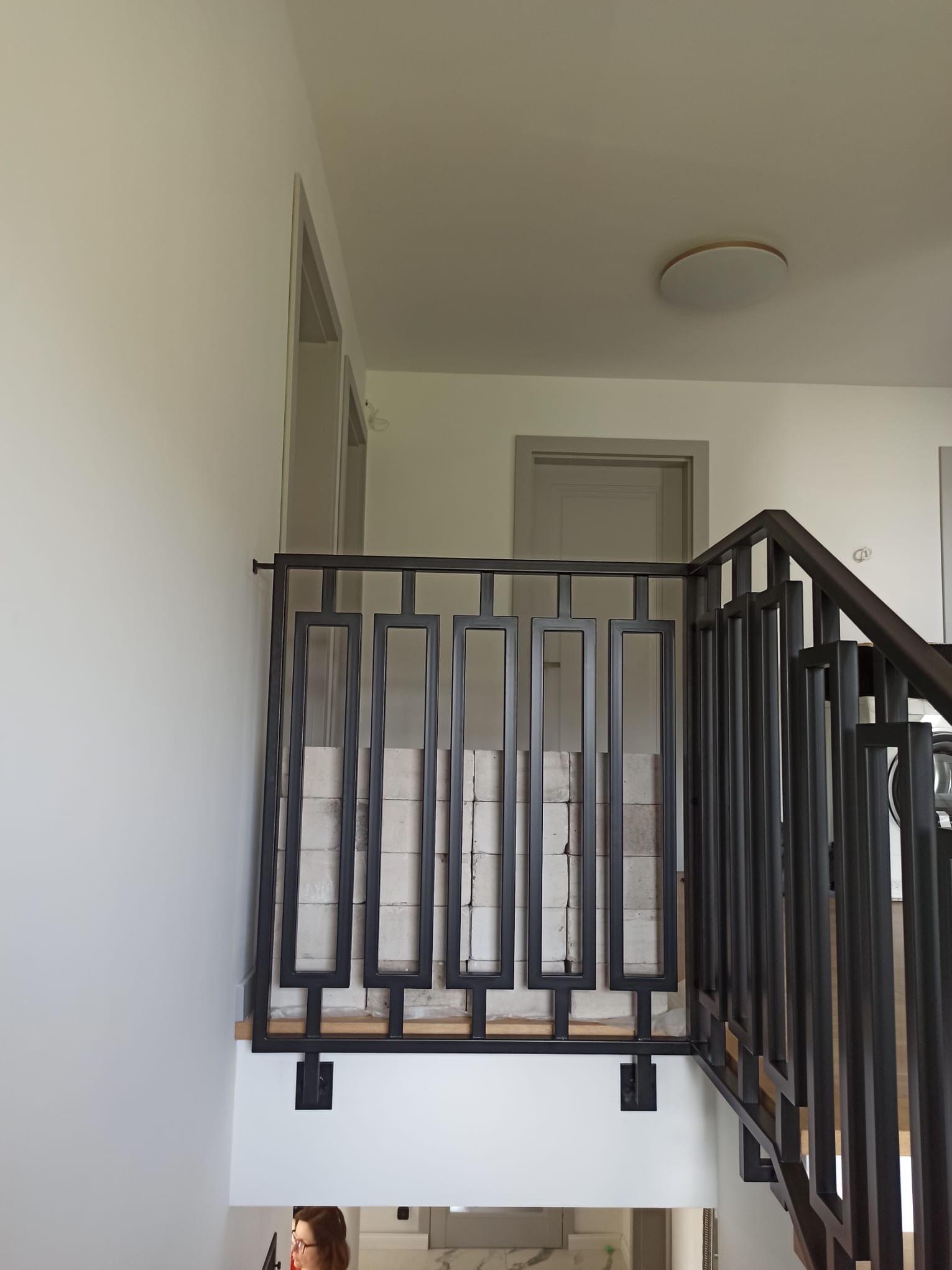 Metalowa balustrada schodowa w nowoczesnym stylu, z geometrycznym wzorem. Widoczne jasne ściany i drzwi w tle. Ciemna kolorystyka balustrady kontrastuje z bielą otoczenia.