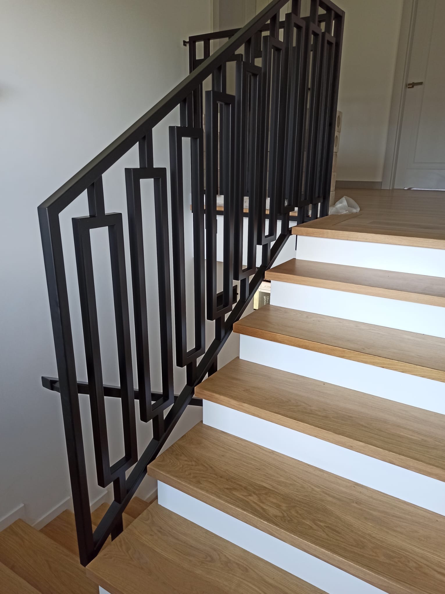 Nowoczesna, czarna poręcz schodowa z geometrycznym wzorem, zamontowana przy drewnianych schodach z białymi podstopnicami, widok z dołu, minimalistyczny design.