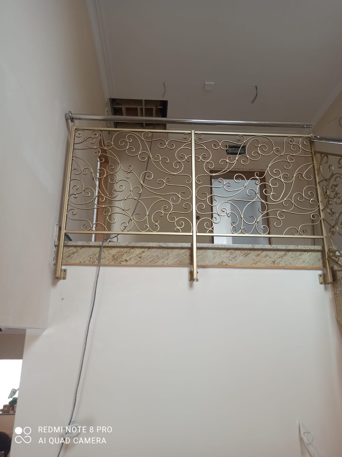 Złota balustrada metalowa z dekoracyjnymi wzorami na tle jasnej ściany i marmurowego parapetu. Widok z dołu na balustradę schodową wewnątrz budynku.