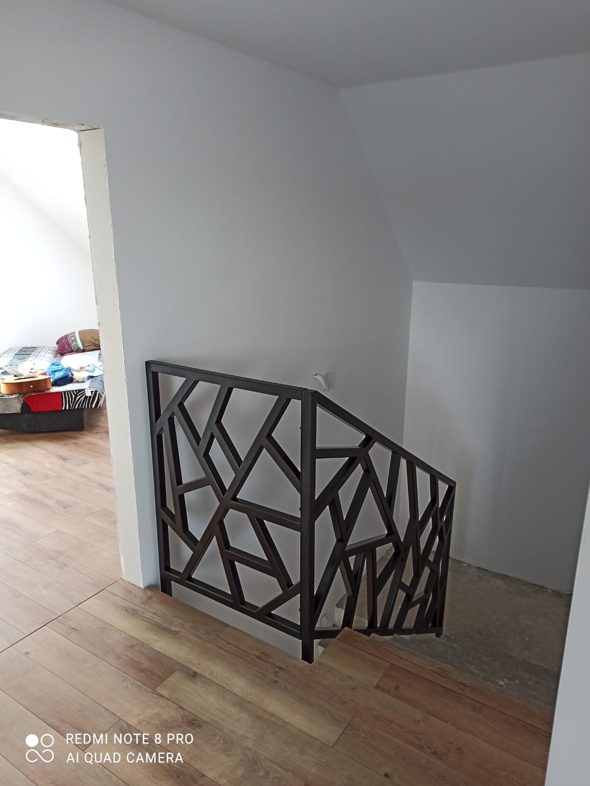 Nowoczesna, geometryczna balustrada metalowa w kolorze ciemnym, zamontowana przy schodach w jasnym wnętrzu domu. Widok z góry na drewnianą podłogę.