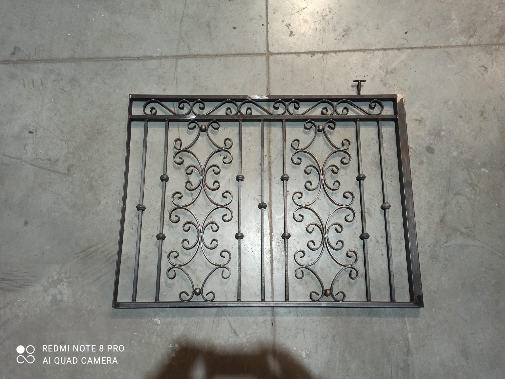 Metalowa krata z dekoracyjnymi, kutymi elementami leży na szarej, betonowej podłodze. Widoczne spawane łączenia i niedomalowane fragmenty.