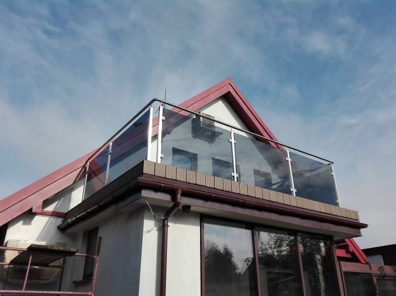 Szklana balustrada balkonowa z elementami ze stali nierdzewnej na tle błękitnego nieba i dachu domu z czerwonej dachówki. Widok z dołu.