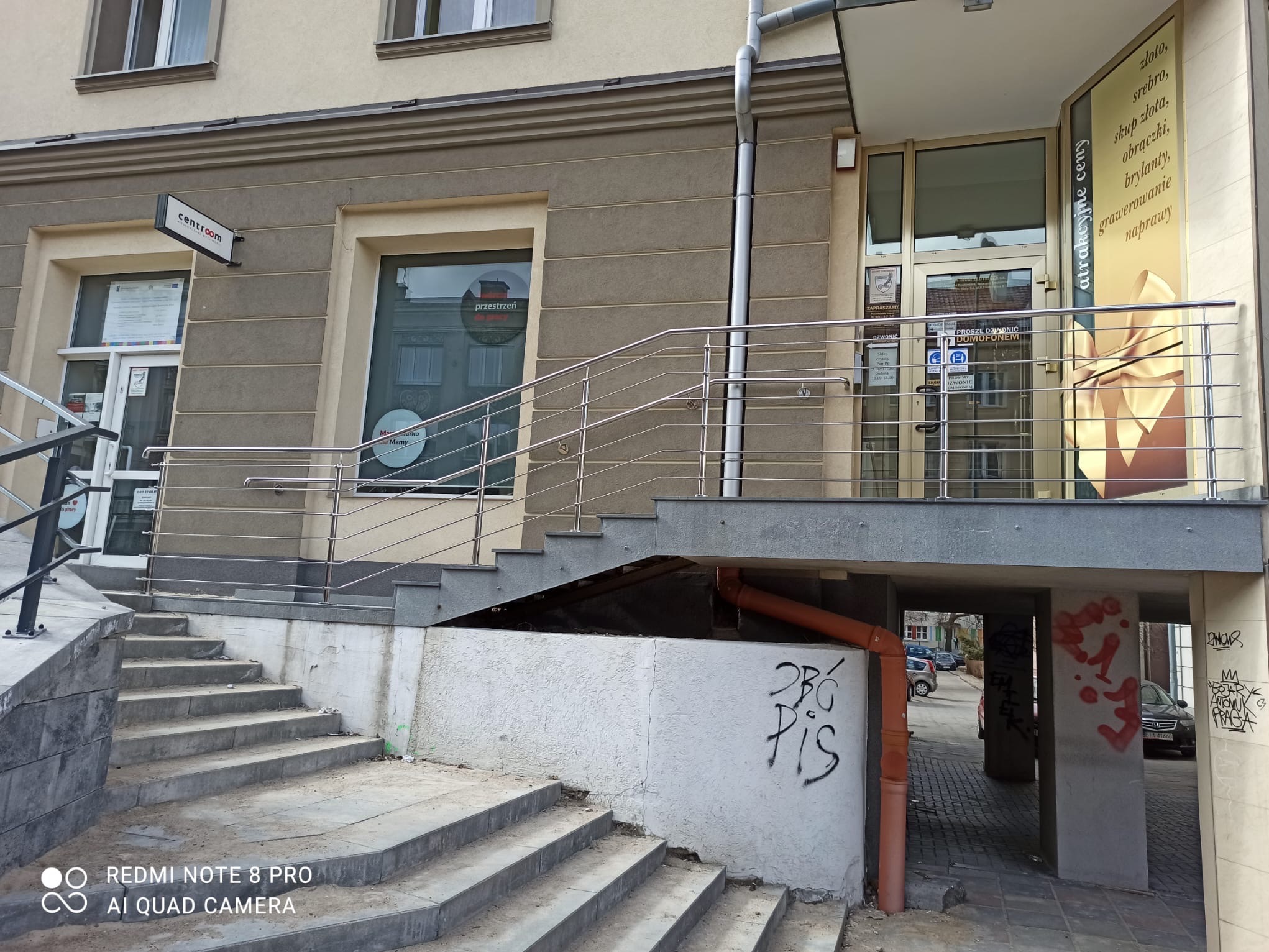 Zewnętrzne schody z metalową balustradą prowadzące do budynku z reklamą usług jubilerskich. Widoczny fragment elewacji i graffiti na ścianie.