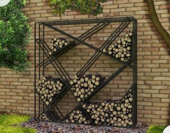 Metalowy regał na drewno kominkowe o geometrycznym wzorze, ustawiony przy ceglanym murze w ogrodzie, z widocznym trawnikiem i drzewem.