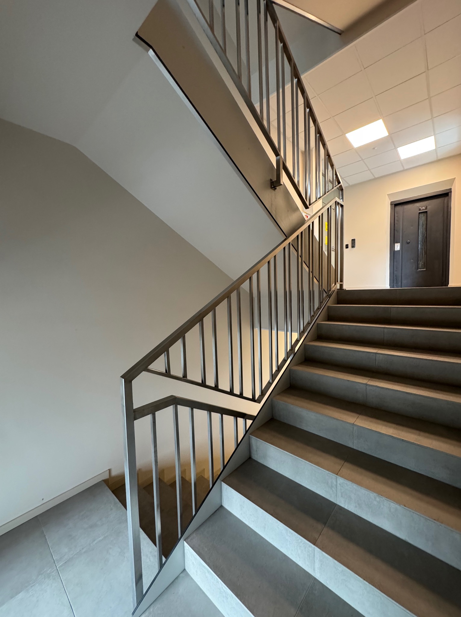 Wewnętrzne schody z metalową balustradą o minimalistycznym designie, prowadzące na wyższe piętro z drzwiami w tle. Widoczne płytki na schodach i podłodze.