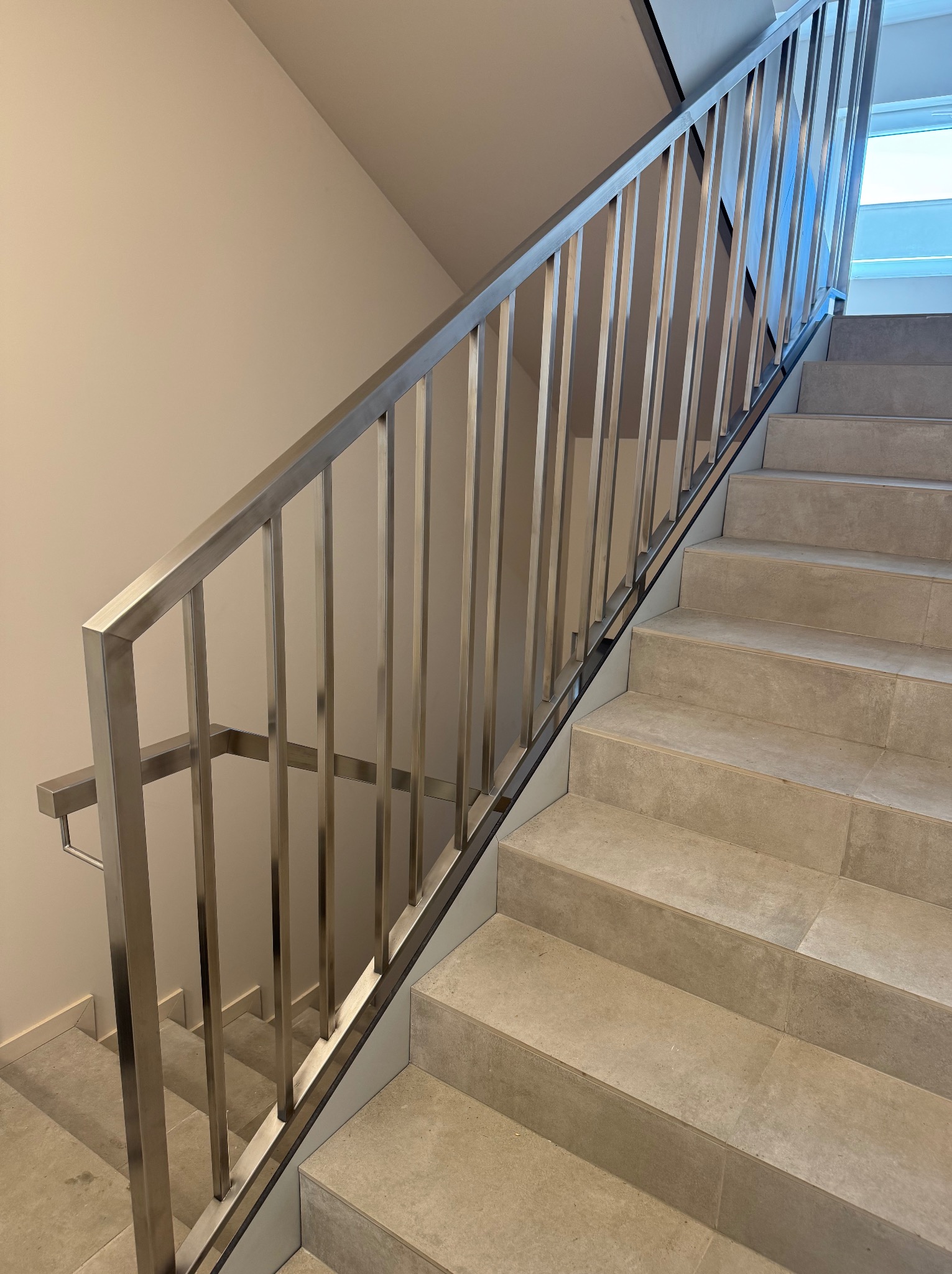 Stalowa poręcz schodowa z pionowymi szczeblami, widok z dołu, jasne płytki na schodach, minimalistyczny design, neutralne tło ściany, światło dzienne.