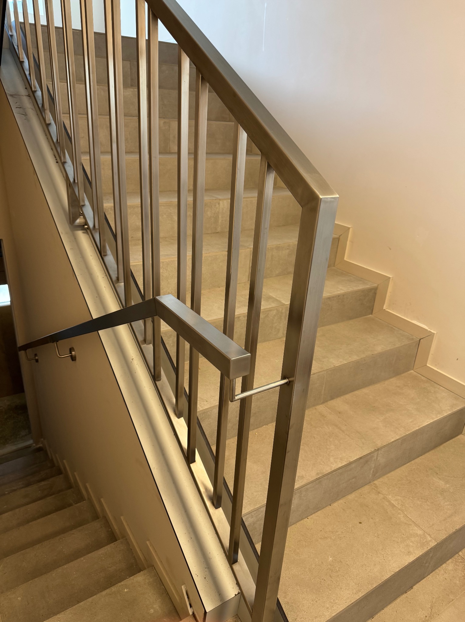 Metalowa poręcz schodowa z pionowymi szczeblami, widok z góry na jasne, wyłożone płytkami schody w budynku. Minimalistyczny design.