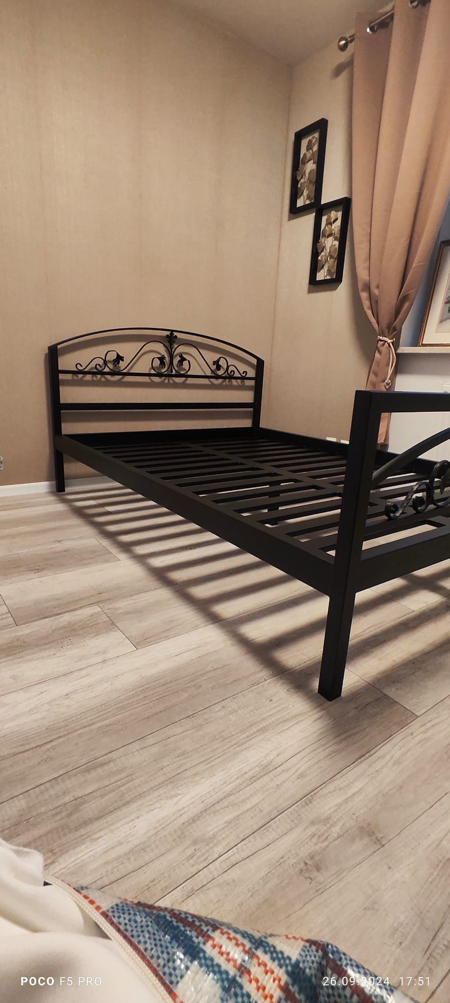 Czarna, metalowa rama łóżka z dekoracyjnym zagłówkiem, widoczna na tle beżowej ściany i drewnianej podłogi. Uwagę przyciąga ornamentyka zagłówka i geometryczny wzór ramy.