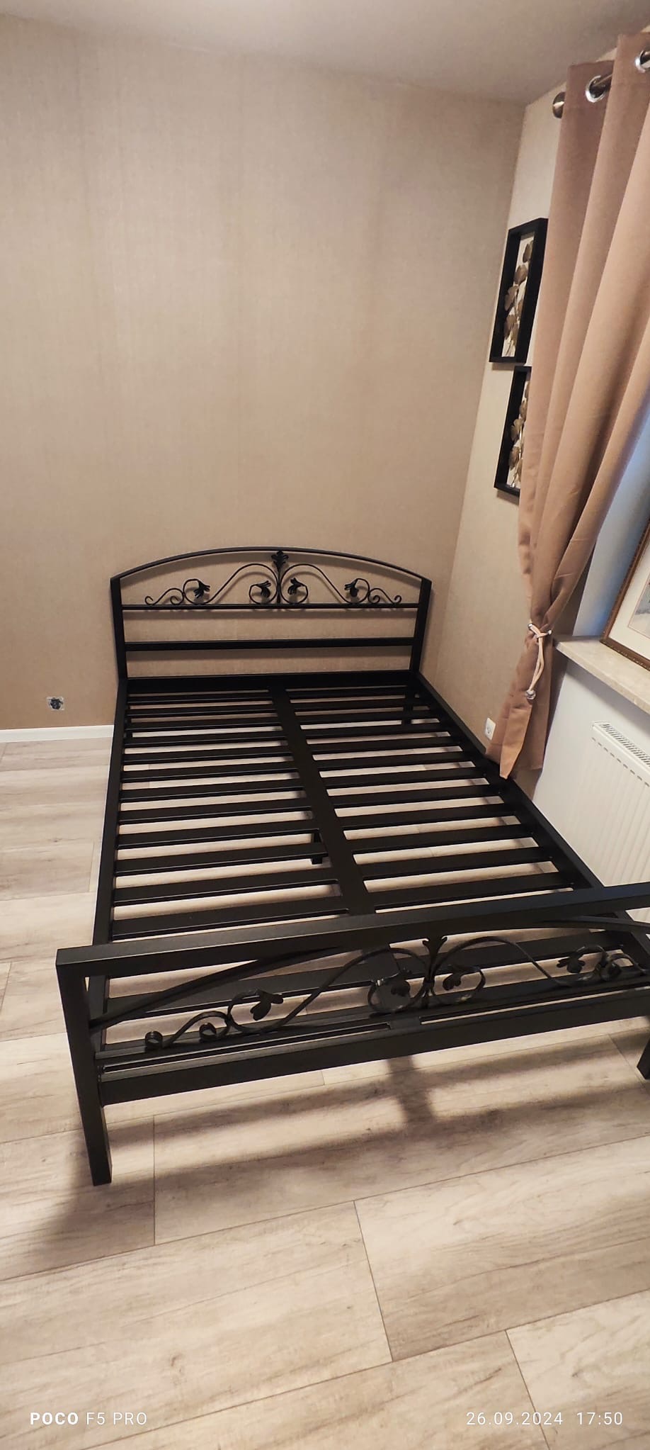 Czarna, metalowa rama łóżka z dekoracyjnymi elementami w sypialni, widok z perspektywy. W tle ściana w odcieniach beżu, zasłona i fragment okna.